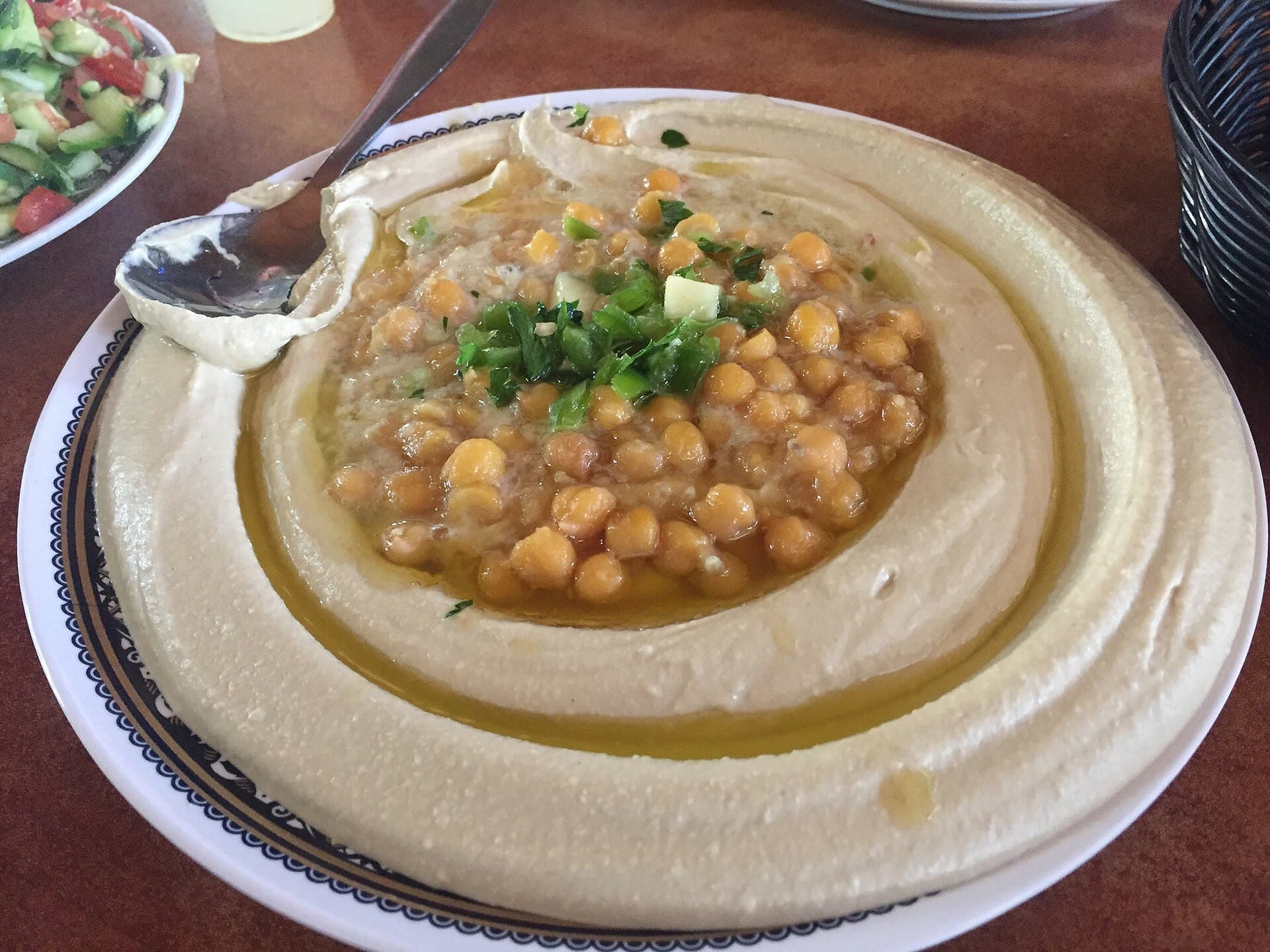 A plate of hummus in a restaurant in Israel, swirled flat with a well of olive oil, whole chickpeas and parsley in the middle, a spoon on the rim and a salad beside it.